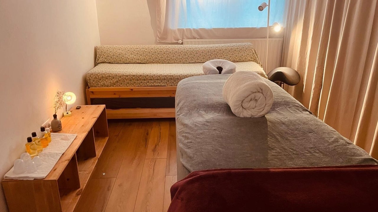 De behandelkamer van Mellow Club: een massagetafel met een opgerolde handdoek, een bijzettafel met olie en een kaarsje, gordijnen voor het raam