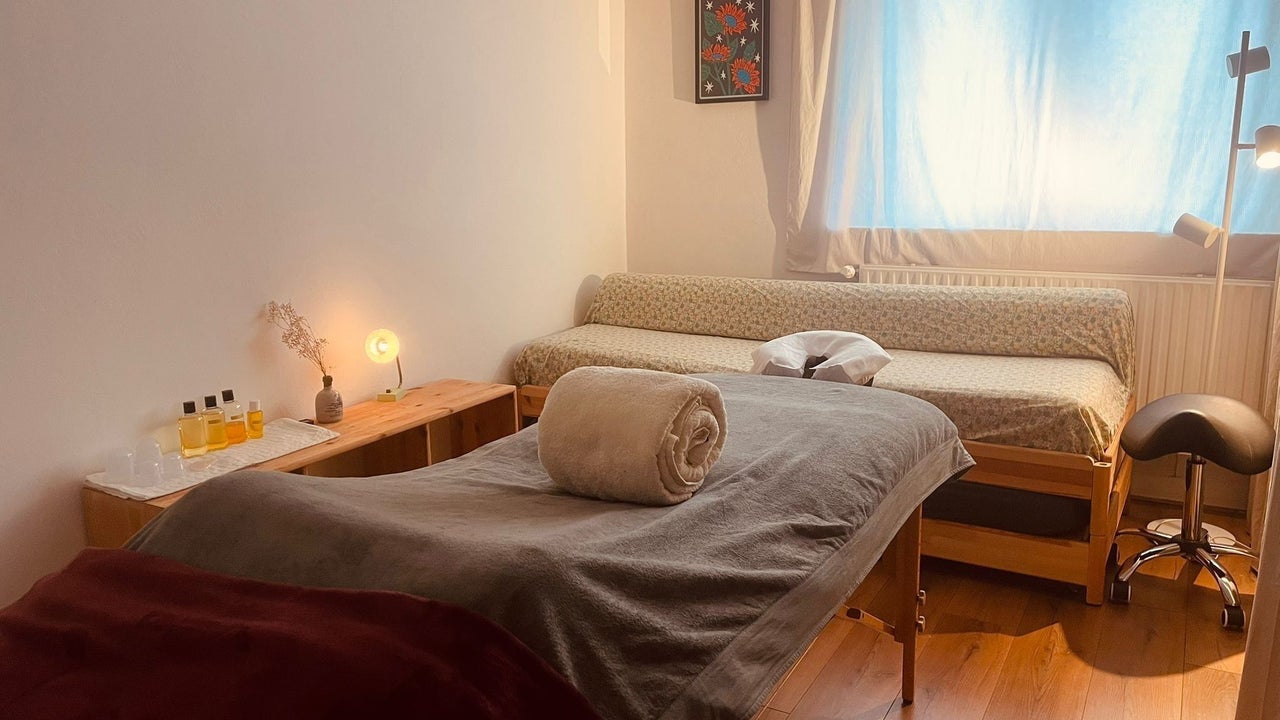 De behandelkamer met de massagetafel, een bank met kussens, een zadelkruk en een raam met gordijnen