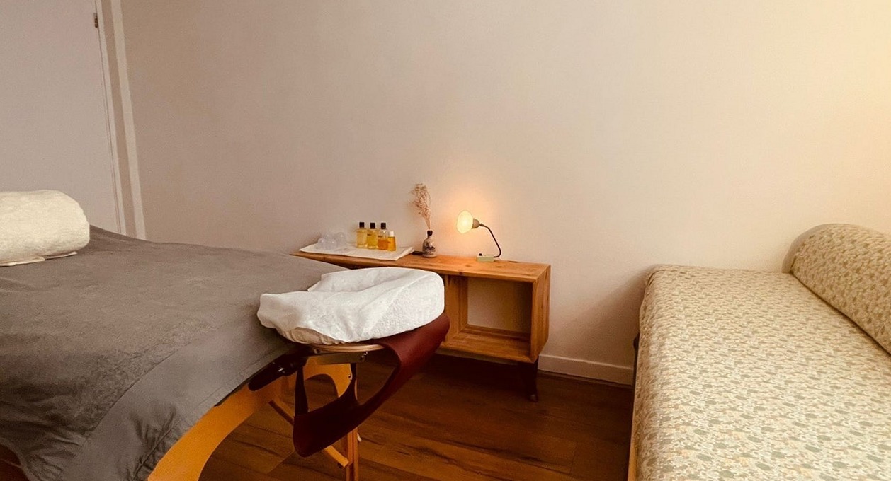Het hoofdeinde van de massagetafel met de hoofdsteun, een bijzettafel met olie en een kaarsje, en een bank met bloemetjesstof